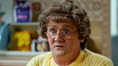 BBC Brendan O'Carroll as Agnes Brown, wearing a yellow cardigan on the show's kitchen set