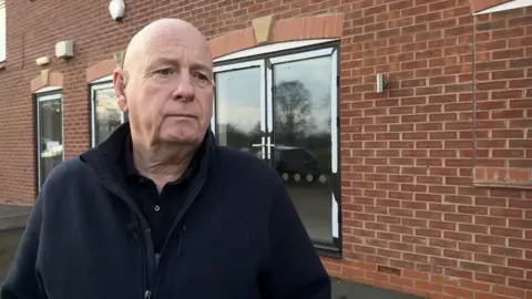 Head and shoulders shot of Steve Lowbridge with a brick house and patio doors in the background. Steve is wearing a dark fleece and dark polo shirt underneath