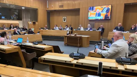 Amy Holmes/BBC A council-style chamber with wooden paneling, where people are seated around a large U‑shaped arrangement of desks. Microphones, laptops, and papers are visible throughout the room, and a screen at the front displays a live feed of the meeting.