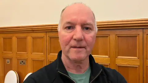 টনি ডোহার্টি একটি কালো জাম্পারের নীচে একটি সবুজ শার্ট পরে আছেন। তার মাথায় বাদামী চুলের দাগ রয়েছে। এর পিছনে দেওয়ালে একটি কাঠের প্যানেল রয়েছে। সেখানে দুটি ক্রিমি চেয়ার বসে আছে। দেয়ালে হালকা সাদা রং করা হয়েছে।