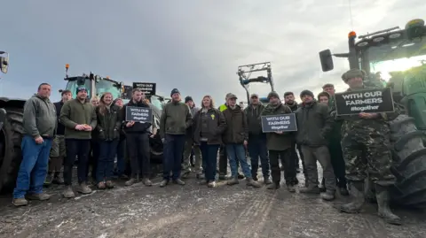 21 farmers stand in front of three tractors. Four of them are holding signs that say "WITH OUR FARMERS #together" written in white block capitals on a black background. 