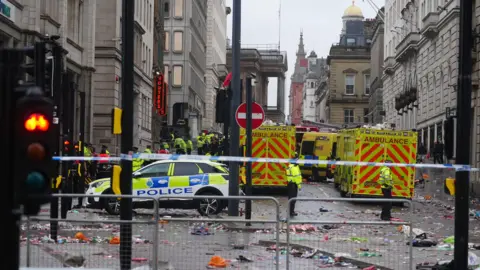 PA Media Police and emergency personnel dealing with an incident near the Liver Building in Liverpool during the Premier League winners' parade. There are at least three ambulances and a police car lying behind police tape. There is much detritus lying on the road.