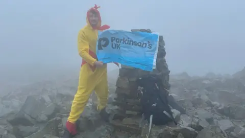 Luke Woodward Luke unfurls a Parkinson's UK flag at the top of the mountain. The surface is rocky, and there is thick fog.