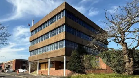 A large four-storey building with alternate layers of brown concrete and glass windows with four white support pillars on the ground floor and a road running at the front.