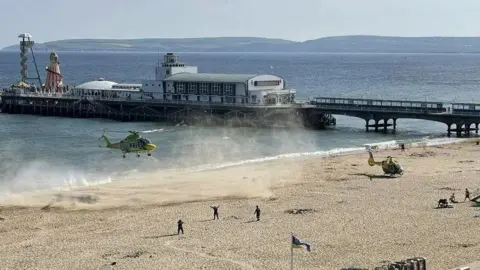 @buhalis Two air ambulances landing on Bournemouth beach with the sea and pier in the background. The beach is empty apart from several emergency service personnel