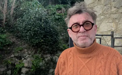 Jim Madden wearing thick framed circular glasses and an orange knitted turtleneck jumper, with a stone wall and bushes in the background.