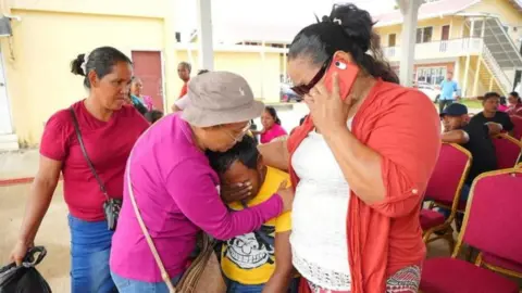 Three women comfort a person who shields their eyes with their hands as they are crying. One of the women is holding a mobile phone to her ear. Another, wearing a pink long-sleeved T-shirt, is placing her arms around the crying person. 