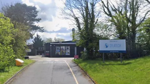 Google The entrance to the Sylatech site, viewed from an access road. A tarmac road runs towards a dark-coloured industrial building. White road markings are visible near the entrance, and there are grassy verges on either side.
To the right, a large white and blue sign reads: WELCOME TO SYLATECH.