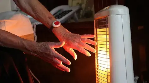 PA Media Close up shot of someone warming their hands in front of a glowing electric heater. A telephone and a family photograph can be seen in the background