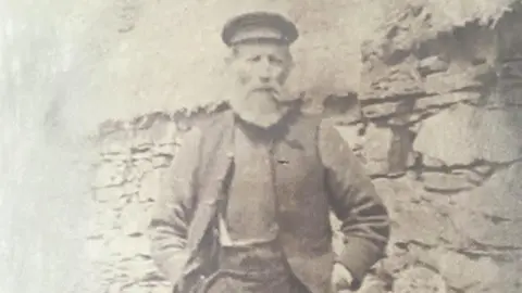 Quirk family An old photo of an older man with a beard smoking a pipe standing outside a stone built thatched cottage.
