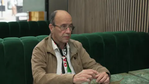 Jonathan Fluxman in a white shirt, Palestine flag scarf, and brown jacket.