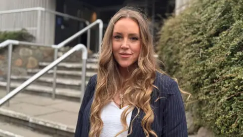 Lindsay Love wears a pin stripe suit jacket over a white top. She has long, wavy brown hair and is standing outside Aberdeenshire Council's office.