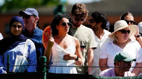 PA Several group opinionated successful a crowd astatine Wimbledon successful nan heat. They are dressed successful summertime clothes, often successful caps and sunhats. The female successful nan centre of nan image is utilizing a instrumentality and wearing sunglasses.