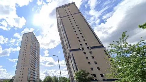 The view from the ground to the top of two tower blocks standing side by side on a sunny day, with just a few fluffy clouds in the sky.
