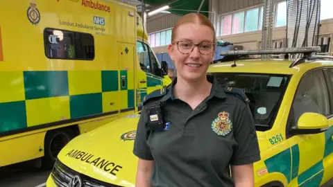 Ella Johnson is wearing a dark green paramedic uniform, and standing with her hands in her pockets and smiling in front of two ambulance vehicles. 