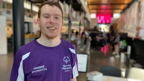 Jon Wright/BBC Freddie Peecock is standing in the foreground of the Beyond Labels exhibition. He has short mousy brown coloured hair and is wearing a branded purple Special Olympics Suffolk shirt. He is looking into the camera and smiling