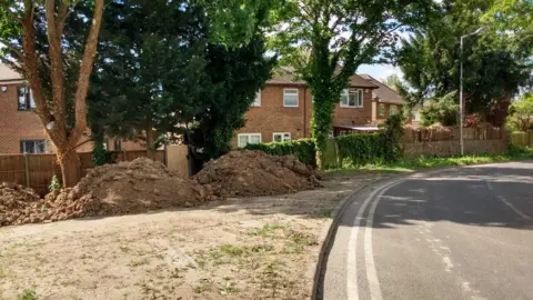 A picture taken aside a road, on the right, and very dry land, with three large piles of dug-up soil, on the left. There are sprigs of grass sprouting from the land. There are homes, which are fenced off, behind the land. 