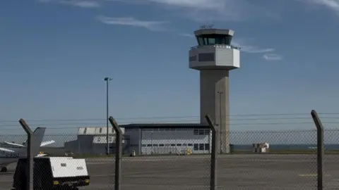 The Air Traffic Control Tower, an industrial looking tower with 360 windows at the top, you can see a fence in front of the tower.