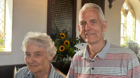 Luke Deal/BBC A man and a woman standing inside a church. They are both smiling. The woman has short grey hair and wears a striped blue and white blouse. The man has short grey hair and also wears a polo top that has grey, white and red stripes on it.
