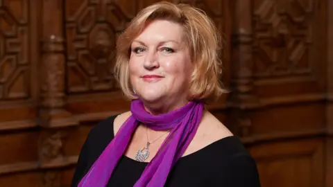Supplied Liz Ward smiling slightly and looking down the camera. She is wearing a black top and a vibrant purple scarf tied around her neck, as well as a silver, glittering necklace. She has auburn hair, cut into a wavy bob. She is standing in front of a wall that is panelled with ornately carved, dark wood.