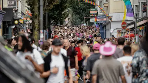 Thousands of people walking up St James' Street in Brighton which is lined with shops and multicoloured pride flags. People are wearing colourful clothing and pink hats
