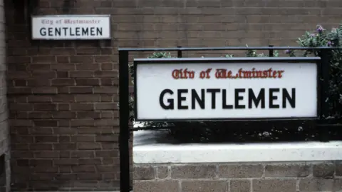 A sign for a public toilet with another smaller identical sign on the wall in the background. The sign is white with "GENTLEMEN" in black lettering and "City of Westminster" in red letters. 