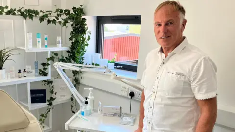 David is standing in a white clinic room with a reclining chair and in front of a table with beauty products which is in front of a window. He is wearing a white shirt and looking into the camera. 