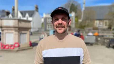 Murdo Mackinnon in a striped T-shirt and wearing a baseball cap. He is standing in a town square on a sunny day. 
