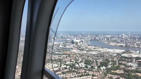 Aerial view through a blimp window showing the River Thames winding through east London, with the O2 Arena visible on the skyline.