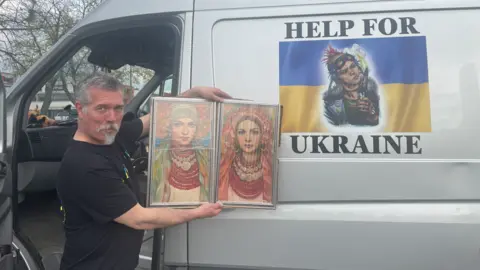 Steve Morris A man wearing a black T-shirt, who has grey hair, is holding images of two women in front of a van. He is standing near an open door and the van includes the words help for Ukraine in black letters.