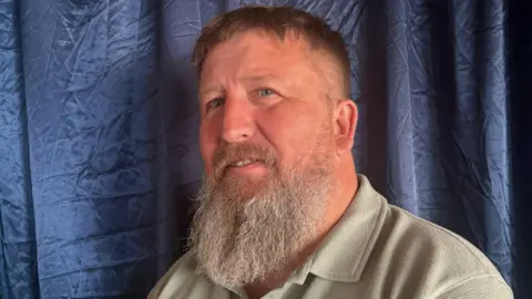 Reform UK's Graham Harrison with long graying beard and wearing a gray-green collared t-shirt stands in front of a creased blue fabric backdrop.