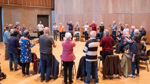 Britten Pears Arts A large group of people stand in a circle, behind their chairs, as they sing. They have their hands lifted in front of them as they sing. They are within a large hall. 