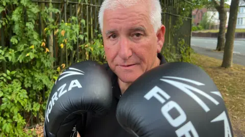 A man in a black T-shirt is standing in front of a tree-lined fence and staring directly at the camera. He is wearing black boxing gloves which are raised to his chin.