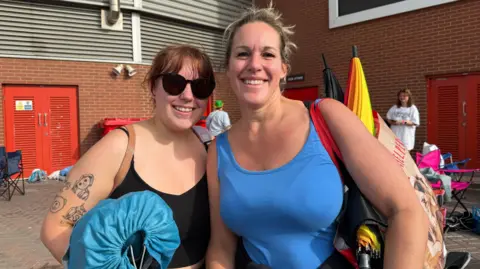 Amy and daughter pose for a picture. They are in summer tops and carry bags and sleeping bags.