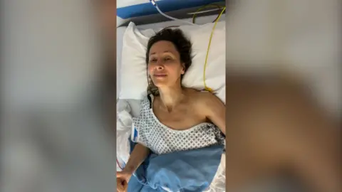 A woman with brown hair in a hospital bed with a gown covering one shoulder