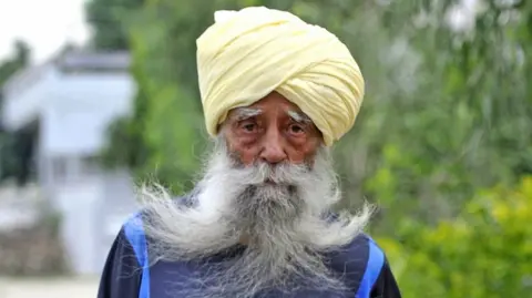 A man in a turban with a long grey beard looks at the camera
