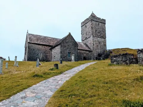 Mag Anderson An old grey stone church with a couple of old grey headstones on a hill with yellowed green grass. A rocky path leads down from the hill to the viewer.