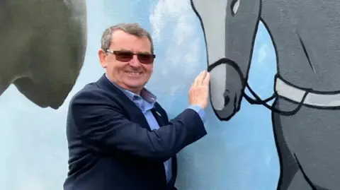Richard Henry with short grey hair and dark glasses, smiling at the camera while wearing a blue shirt and a dark blue jacket. He is touching the nose of a horse depicted on a mural behind him.