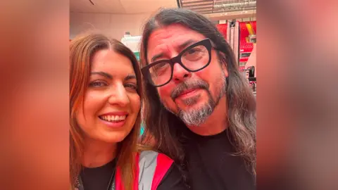 Danielle McNally Danielle McNally has red hair and wears a hi-vis jacket. She is smiling stood next to a man with long dark hair and a silver beard and glasses, Dave Grohl.