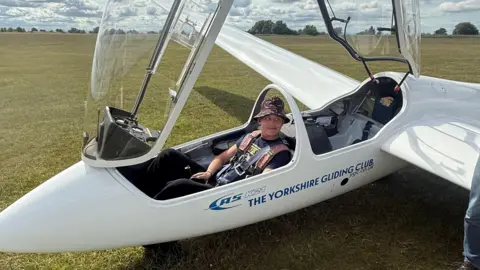 Kamila Nowicka A teenage boy is sitting in the front seat of a glider on the ground which has its clear cockpit raised. 
