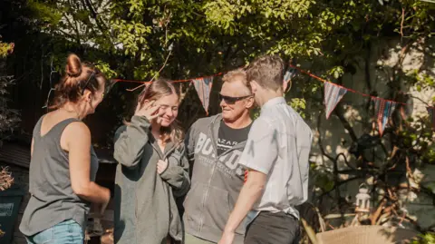 Fine Rolling Media Mark returning home after his second stem cell transplant. He is in black sunglasses and a grey hoodie and is stood next to his daughter, also in grey hoodie with brown hair, his wife, who is in a grey vest top and is facing the wrong way and his son, who is in a light grey shirt and also has his back to camera