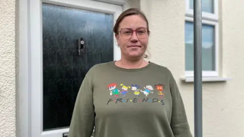 BBC A woman with brown hair, wearing glasses with a diamond stud under her nose. She's wearing a green jumper with the word Friends on it and the characters from the Rugrats. 