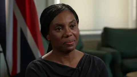 Kemi Badenoch, wearing a black dress, sits in a room. There are green armchairs and a union flag visible in the background. 