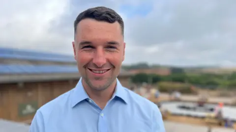 A man in a light blue shirt smiles at the camera while standing outside Occombe Farm in the sun