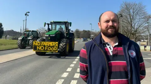 A man standing on the side of the road with a his hands behind his back.  There is a tractor on the road to the left and is has a black and yellow sign that reads "no farmers, no food". The sky is blue in the background