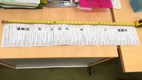 SIMON DEDMAN/BBC A long sheet of paper sits on wood-topped tables, with various boxes printed on it. A yellow tape measure is extended parallel to the paper.