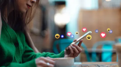 Getty Images A woman looking at a phone with emojis representing social media