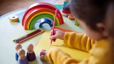 A child wearing a bright yellow jumper draws with a pink colouring pencil on a yellow piece of paper. There is a wooden rainbow and more pencils on the desk.