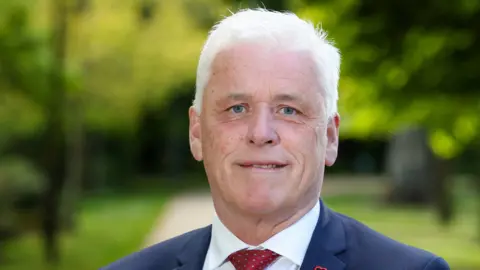 British Heart Foundation A close-up photo of Fearghal McKinney standing in a park on a sunny day. He has short, white hair and blue eyes. He is wearing a navy suit jacket, a white shirt and a red patterned tie.
