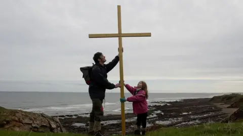 Pete Coppola A small girl wearing a pink jacket and with long brown hair is holding a cross and looking up at it. Next to her is a man who is also holding the cross. They are standing at the coast with the sea behind them